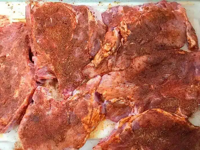 Several raw pork chops covered in a reddish dry spice rub, arranged on a white surface, ready to become the best baked pork chops for your next meal.