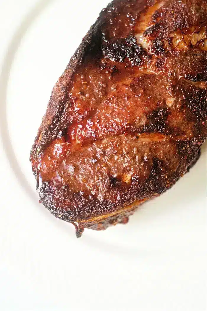 A close-up image of a well-cooked steak, showcasing a crispy, seared crust and juicy texture on a white plate. The meat appears to be seasoned with spices and herbs, similar to an air fryer pork roast, giving it a flavorful, caramelized exterior.