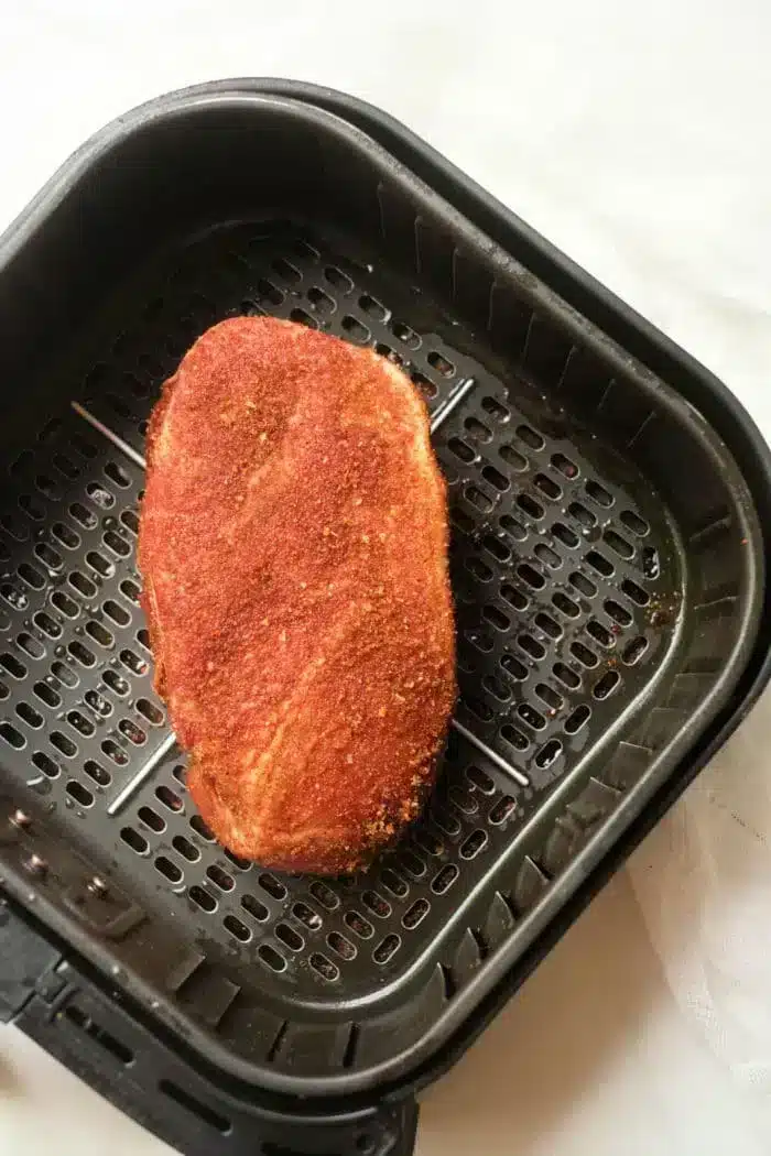 A piece of seasoned food, likely air fryer pork roast or tofu, sits in a black air fryer basket. The surface is coated with a reddish spice mix, and the basket has a perforated design for air circulation. The surrounding area appears to be a white surface.