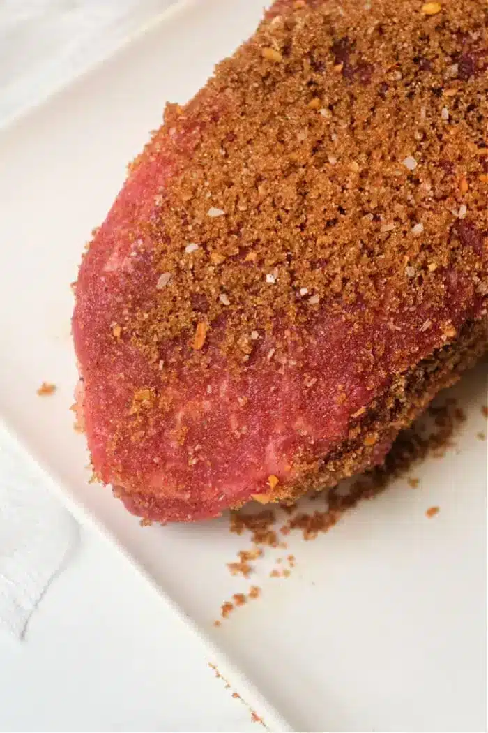 A close-up of a piece of raw meat, likely air fryer pork roast, coated with a brown seasoning rub, placed on a white surface. The seasoning appears coarse and covers the entire visible part of the meat, with some granules scattered on the surface below.