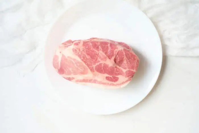 A raw cut of marbled meat is placed on a white plate, ready to become a succulent air fryer pork roast. The plate rests on a light-colored surface, and the background appears to be a white textured fabric.