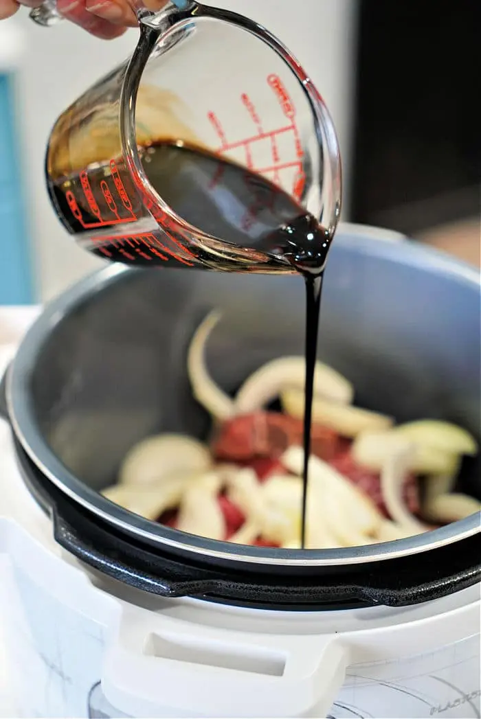 A hand is pouring a dark liquid from a measuring cup into an electric cooker containing sliced onions and beef arm roast. The cooker's lid is open, spotlighting the ingredients as they meld into a mouthwatering recipe.