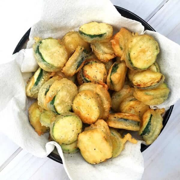 A bowl of crispy salt and pepper zucchini chips rests on a paper towel-lined plate.