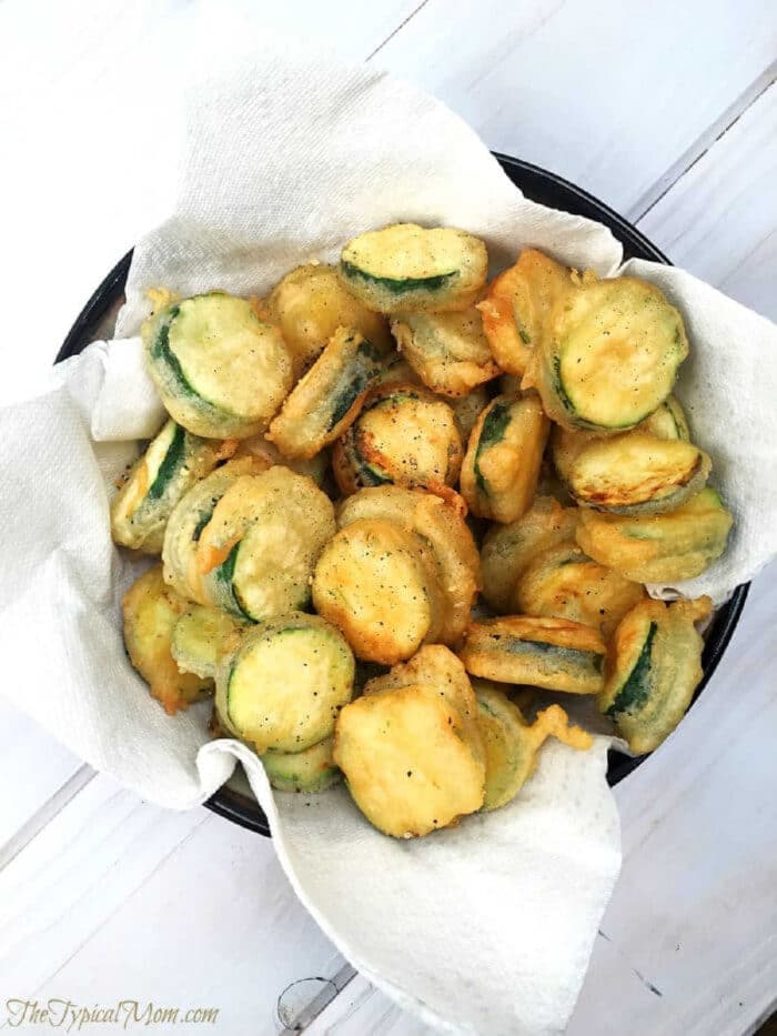 A bowl of crispy salt and pepper zucchini chips rests on a paper towel-lined plate.