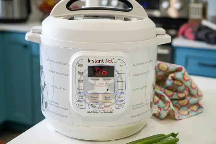 A white Instant Pot with a Star Wars theme sits on the kitchen counter, ready to tackle your favorite beef arm roast recipe. The digital display glows "On." Next to it lie a patterned cloth and some green onions, all against the backdrop of blue kitchen cabinets and a stovetop.