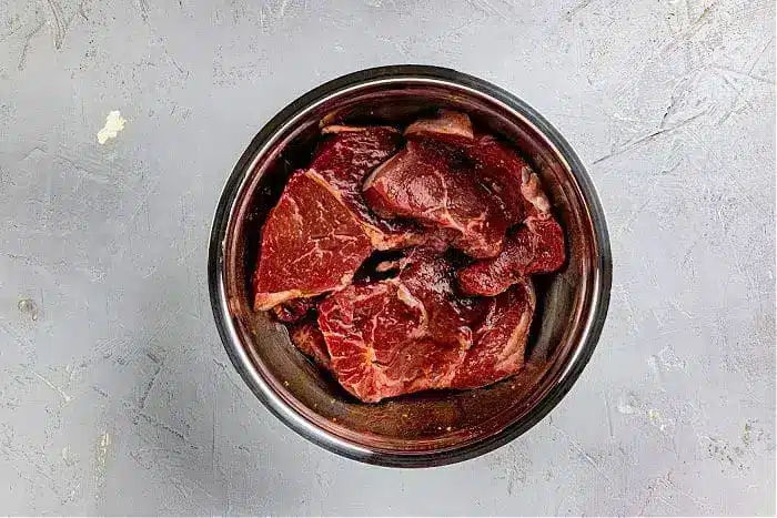 A metal bowl filled with raw pieces of beef, ideal for an Asian steak marinade, sits on a gray textured surface. The beef is marbled and visibly fresh. The bowl is the focal point of the image.