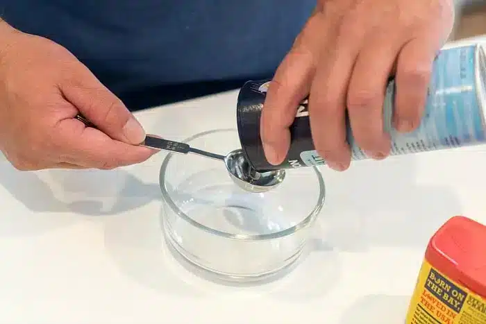 A person carefully measures salt from a red-lidded container into a measuring spoon, poised above a small glass bowl on a white surface, perfect for enhancing the flavors of the best baked pork chops.