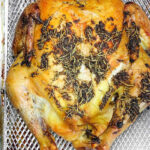 Whole roasted chicken with herbs on its skin sits in a metal basket, likely from an air fryer or oven—perfect for transforming frozen chicken in oven into a delicious, golden-brown meal.