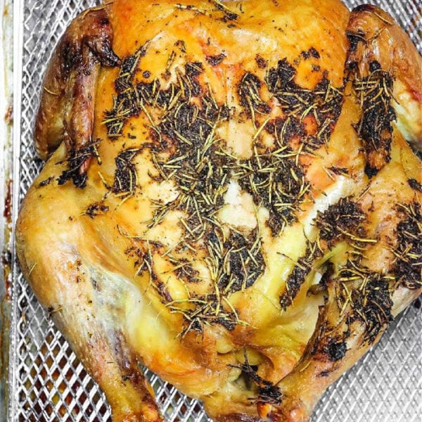 Whole roasted chicken with herbs on its skin sits in a metal basket, likely from an air fryer or oven—perfect for transforming frozen chicken in oven into a delicious, golden-brown meal.