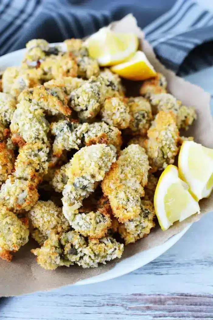 A plate of breaded and fried oyster mushrooms, reminiscent of air fryer fish in texture, is served with lemon wedges. The mushrooms have a golden-brown, crispy crust and are arranged on a paper-lined dish. A gray cloth napkin is partially visible in the background.