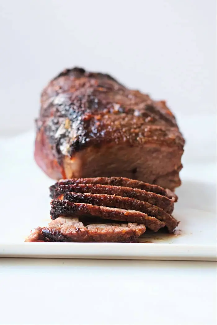 Image of a roasted beef brisket with a dark, crispy crust, sitting on a white cutting board. The brisket is partially sliced, showing its interior texture and juiciness. Prepared using the air fryer roast beef method, the background remains neutral and plain.