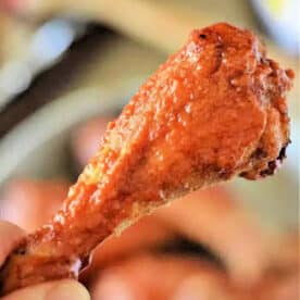 A hand holding a crispy, saucy baked chicken leg, reminiscent of buffalo chicken drumsticks, with a blurred background.