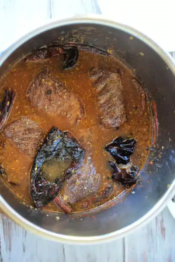 A pot with chunks of beef simmering in a reddish-brown Birria sauce, accented by visible dried chili peppers and spices. The dish, rich in flavor and aroma, is likely being cooked on a stove.