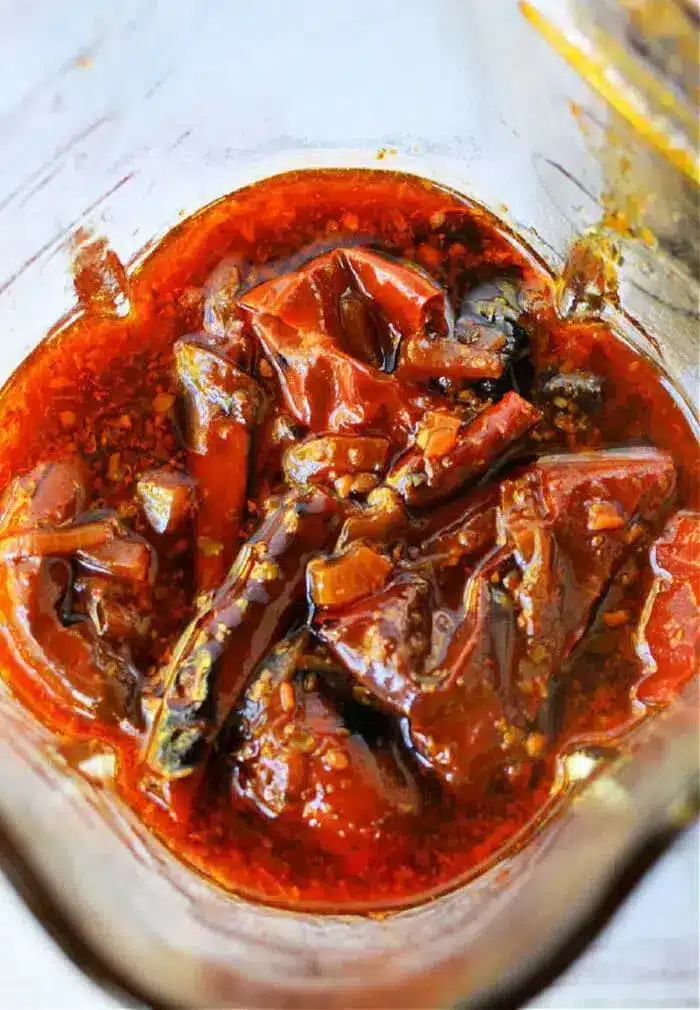 A close-up of dried red chilies soaking in a rich, oily blend of spices and seasoning, reminiscent of a vibrant birria tacos sauce. The marinade is a luscious reddish-brown hue, with the chilies appearing softened and perfectly coated in the flavorful oil.