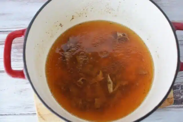 A red-handled pot containing simmering broth reminiscent of a brown gravy for meatloaf recipe with visible meat bones rests on a wooden surface. The rich, brown broth is slightly foamy at the edges.