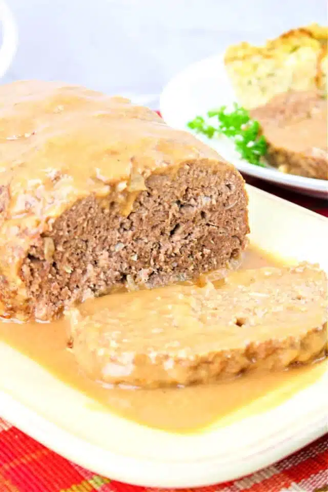 A loaf of meatloaf topped with rich brown gravy is served on a white plate. A slice has been cut from the front, revealing its texture. In the background, there is a plate with side dishes, including peas—a perfect scene for our savory brown gravy for the meatloaf recipe.