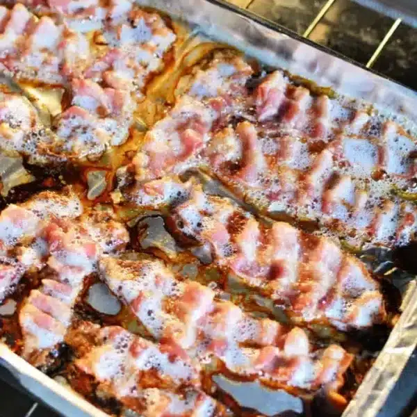 A baking tray lined with aluminum foil holding cooked slices of bacon is placed in an oven.