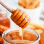 A honey dipper drizzling hot honey sauce into a glass jar, with blurred sticks and a jar in the background.