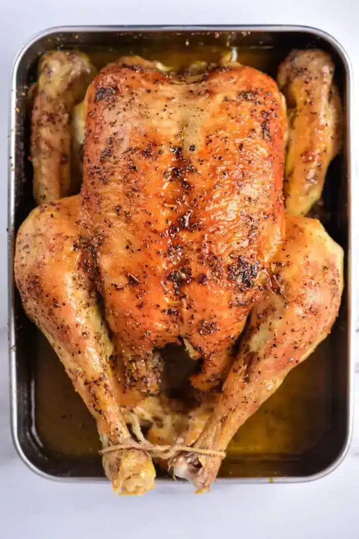 A roasted whole chicken in a baking tray, seasoned with herbs and spices, with tied legs. The skin is golden brown and crispy, resting in a pool of juices. Wondering how long to bake a whole chicken at 350? Ensure perfect results by timing your bake for about 1.5 to 2 hours.