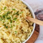 A bowl of perfectly cooked orzo pasta garnished with fresh parsley rests on a wooden board, accompanied by a rustic wooden spoon. Curious about crafting this dish? Discover how to cook orzo effortlessly to enjoy at home.