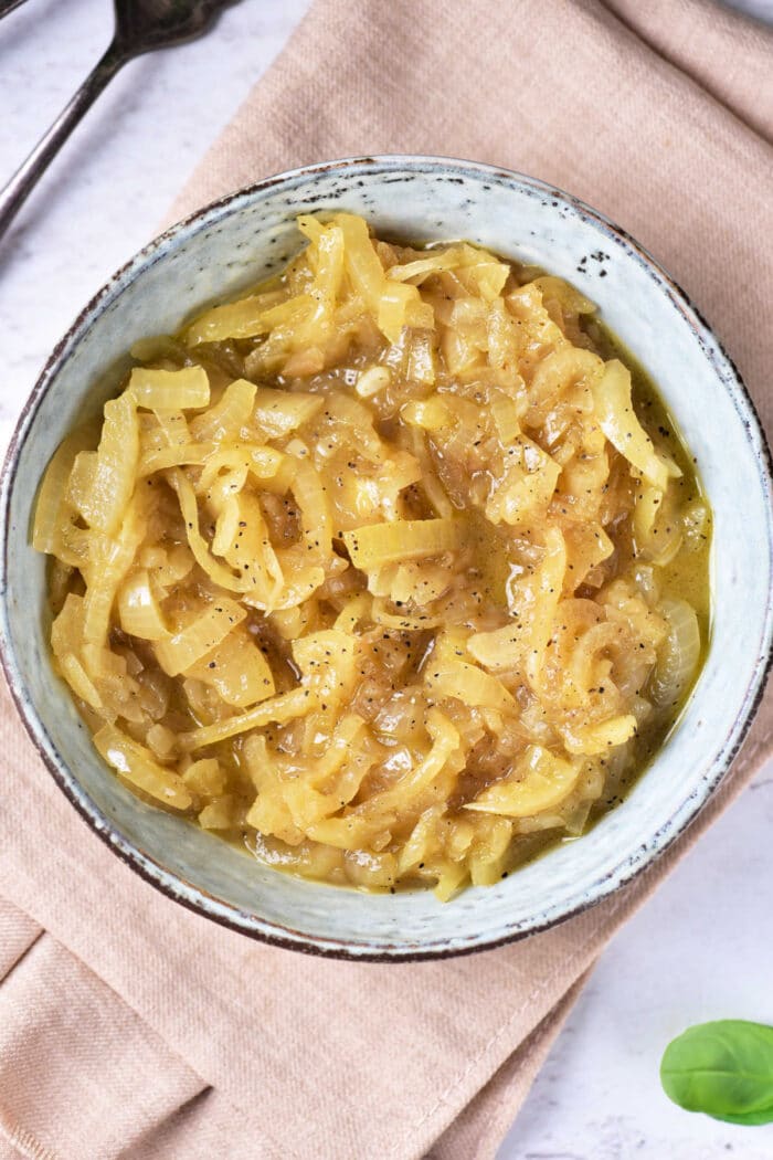 A bowl of onion confit rests on a beige napkin. The onions are golden brown, glossy, and seasoned lightly with black pepper. Cutlery is partially visible in the background.