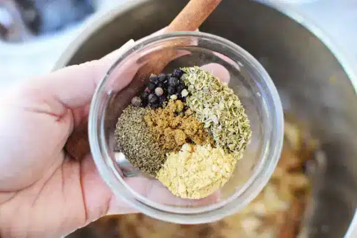 A hand holds a glass bowl of spices perfect for birria tacos sauce, featuring whole black peppercorns, dried oregano, ground cumin, coriander, and ginger. In the background, a wooden spoon awaits to blend these flavors into a savory masterpiece.