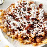 A delightful plate of funnel cake, dusted with powdered sugar and sprinkled with crunchy bacon bits, offers a heavenly taste experience. Perfect for those curious about how to make a funnel cake masterpiece.