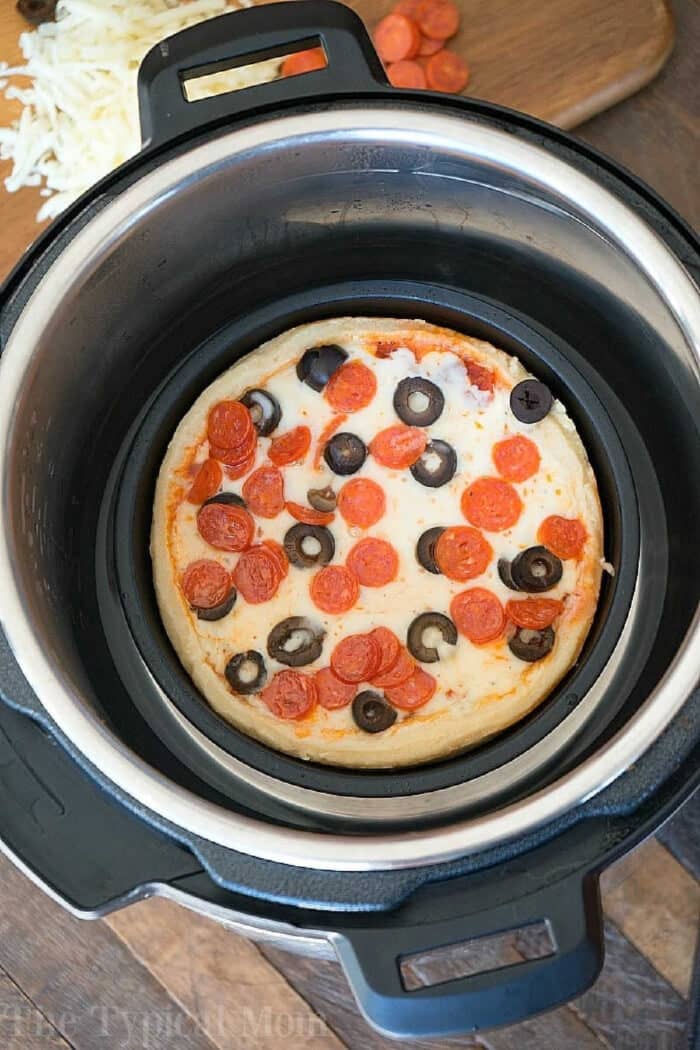Instant pot pizza perfection: a personal-sized pepperoni and olive pizza cooks inside an Instant Pot, with fresh toppings ready on a cutting board.