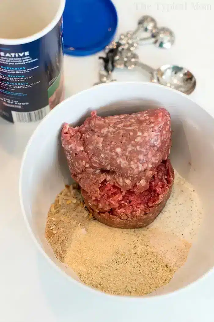 A bowl containing ground meat and various spices ready to become a delicious Ninja Foodi meatloaf. There's a container in the background and a set of measuring spoons.