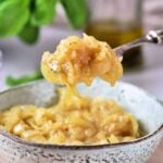 A spoonful of onion confit rests above a speckled ceramic bowl, with a blurred green plant in the background.