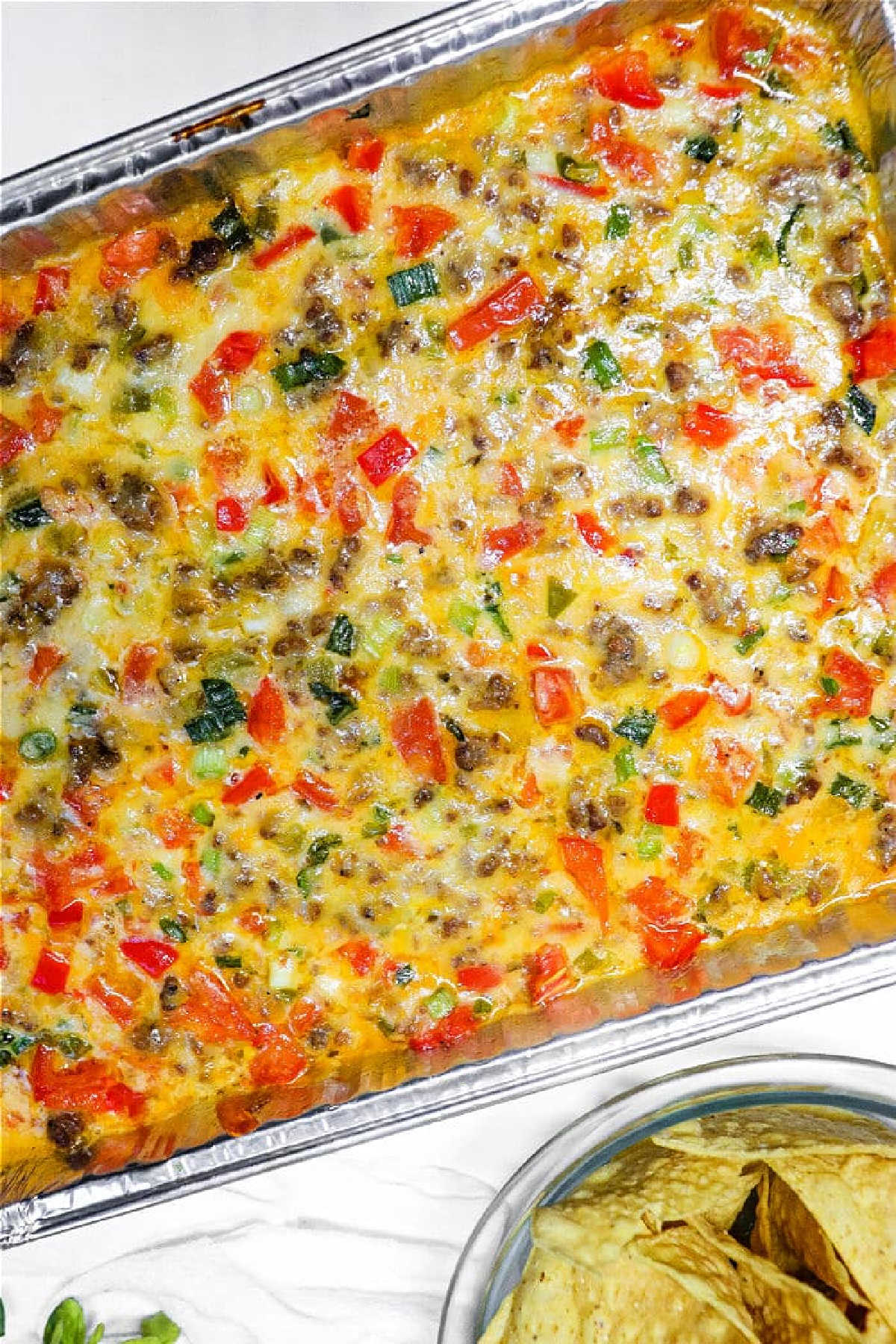 Baked breakfast casserole with sausage, cheese, colorful vegetables, and a hearty queso dip with meat in a foil tray, served next to tortilla chips.