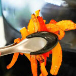 Tongs holding cooked orange and red air fryer peppers in front of an air fryer.