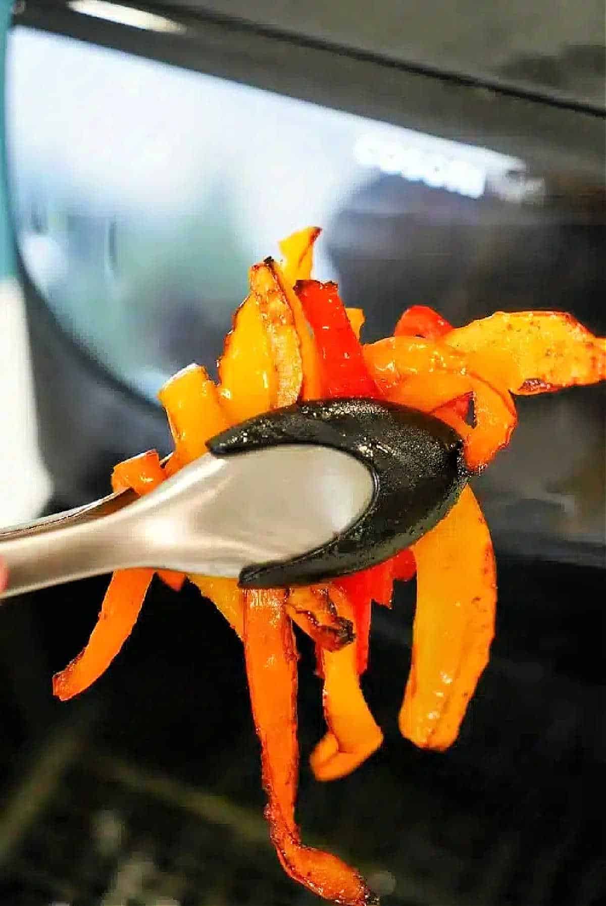 Tongs holding cooked orange and red air fryer peppers in front of an air fryer.