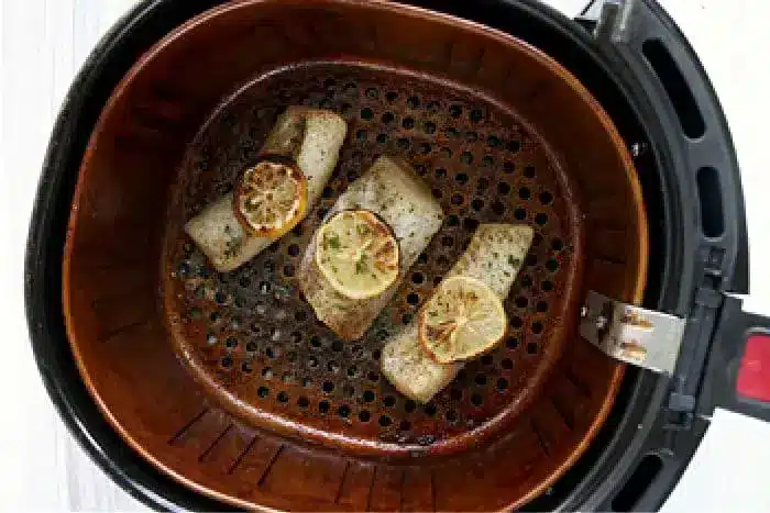 An air fryer basket containing three pieces of seasoned fish fillets topped with lemon slices. The air fryer fish sits in a sleek black exterior unit with a visible latch. The interior of the basket shows some cooking marks and residue, evidence of its recent culinary success.