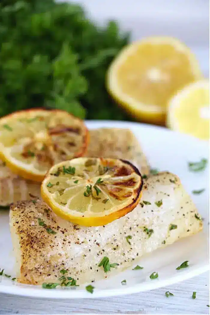 A plate of air fryer fish fillets seasoned with herbs and black pepper, topped with slices of grilled lemon. In the background, there are fresh parsley and halved lemons.