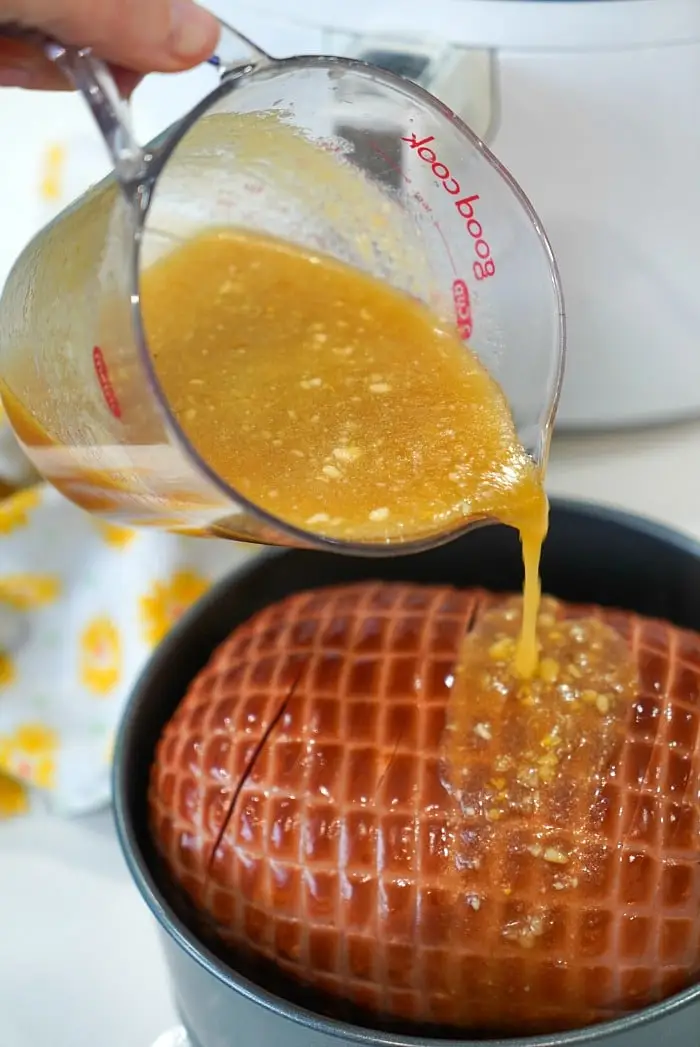 Close-up of a hand pouring a yellowish glaze with small chunks from a glass measuring cup onto an air fryer ham in a black pot. In the background, you can see a white appliance and a yellow and white patterned cloth.