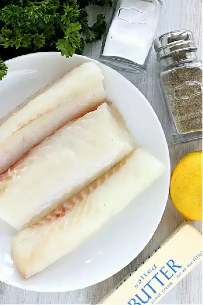 A plate of three raw fish fillets rests on a light wood surface. Surrounding the plate are a small pile of parsley, a salt shaker, a pepper shaker, a lemon, and a stick of salted butter. Perfect for making air fryer fish, these ingredients promise ease and flavor in every bite.