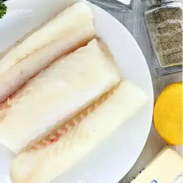 A white plate contains three raw fish fillets, ready for the air fryer. Surrounding the plate are a bunch of parsley, a salt shaker, a pepper shaker, half a lemon, and a stick of salted butter.