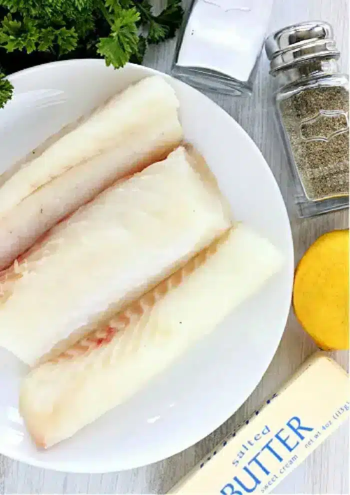 A white plate contains three raw fish fillets, ready for the air fryer. Surrounding the plate are a bunch of parsley, a salt shaker, a pepper shaker, half a lemon, and a stick of salted butter.