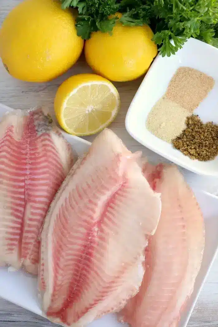 Three raw fish fillets are placed on a white plate, perfect for preparing air fryer fish. Beside the fillets are three whole lemons, one half lemon, a pile of fresh parsley, and a small dish containing brown and white spices. The image is set on a light-colored wooden surface.