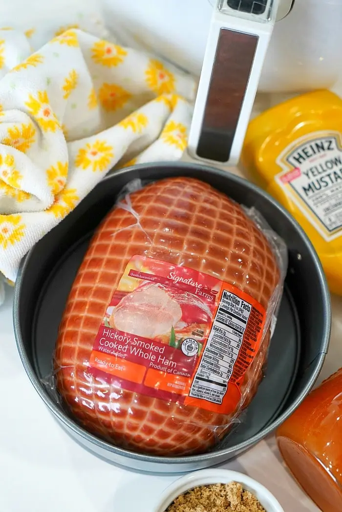 A round pan containing a packaged hickory-smoked cooked whole ham sits on a counter, ready to be transformed into a delicious air fryer ham. Surrounding items include a tub of yellow mustard, a small container of brown sugar, a meat thermometer, and a cloth with a yellow floral pattern.