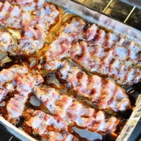 Bacon strips sizzling on a foil-lined baking sheet inside an oven show exactly how to cook bacon in the oven with aluminum foil.