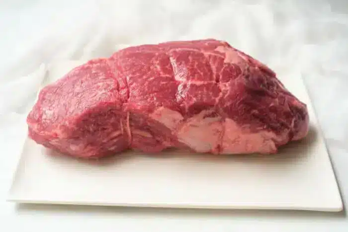 A raw cut of beef placed on a rectangular white ceramic platter, showcasing its visible marbling and fat. The background is light and softly blurred, highlighting the red color and texture of what promises to be a delectable air fryer roast beef.