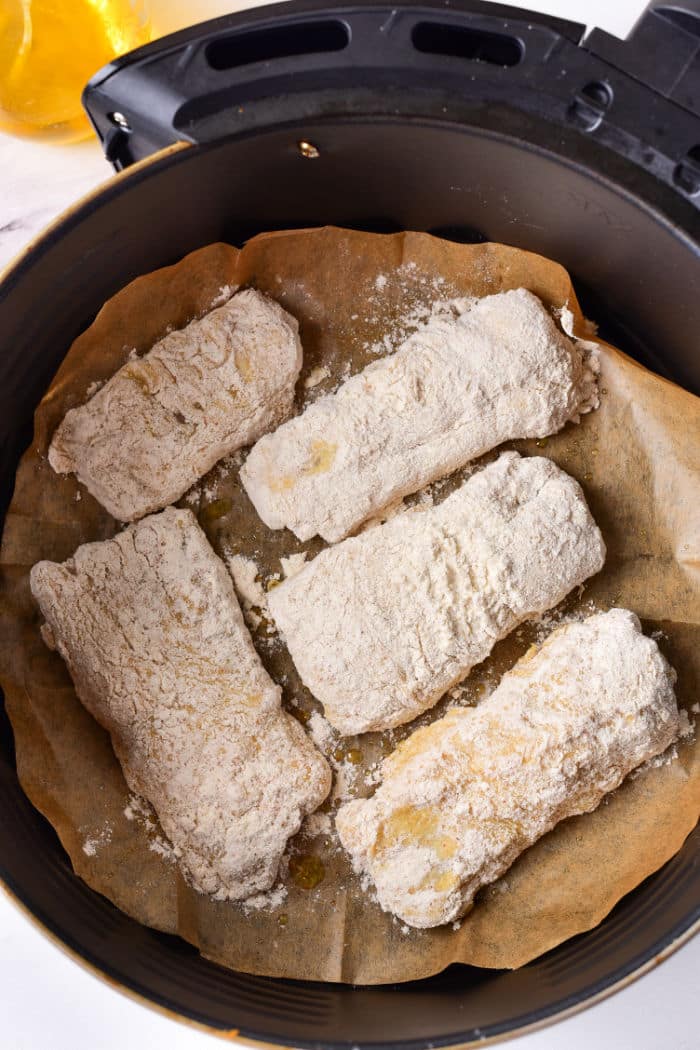 Five pieces of beer battered fish, coated in flour, rest on parchment paper inside an air fryer basket, ready to be cooked to crispy perfection.