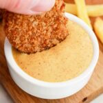 A hand dips a chicken nugget into creamy dipping sauce, reminiscent of those signature flavors youd find when learning how to make Burger King sauces, with crispy fries in the background on a wooden board.