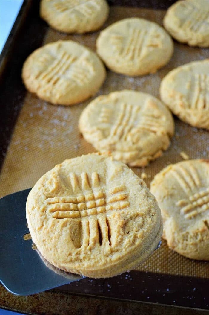 A metal spatula lifts a peanut butter cookie with a crisscross fork pattern from a baking tray, showcasing cookies made with cake mix for an extra soft texture.