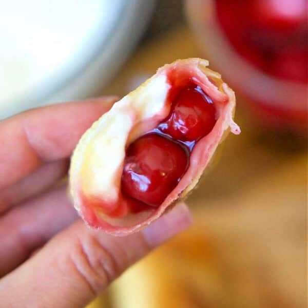 A hand holds a cherry cheesecake egg roll filled with creamy cheese and cherry pie filling, showing the delicious inside.