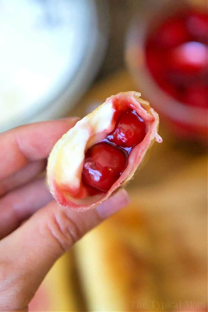 A hand holds a cherry cheesecake egg roll filled with creamy cheese and cherry pie filling, showing the delicious inside.