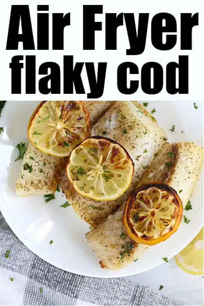 A plate of air fryer fish cod fillets is garnished with slices of lemon and sprigs of parsley. The fish appears to be seasoned with pepper and herbs.