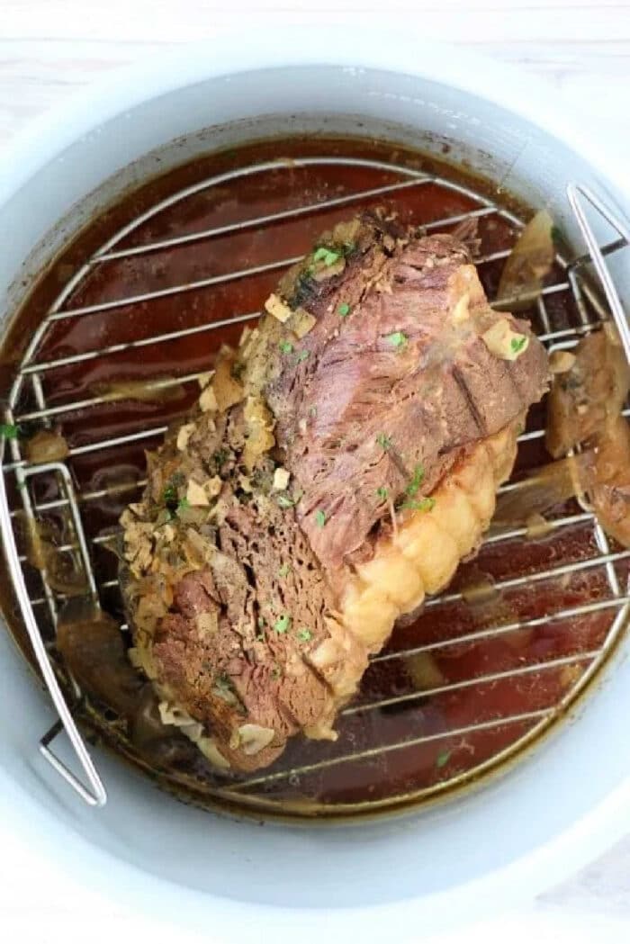 Cooked beef roast topped with herbs and garlic sits on a metal rack inside a pot with broth, perfect for making a tender frozen roast in Instant Pot.